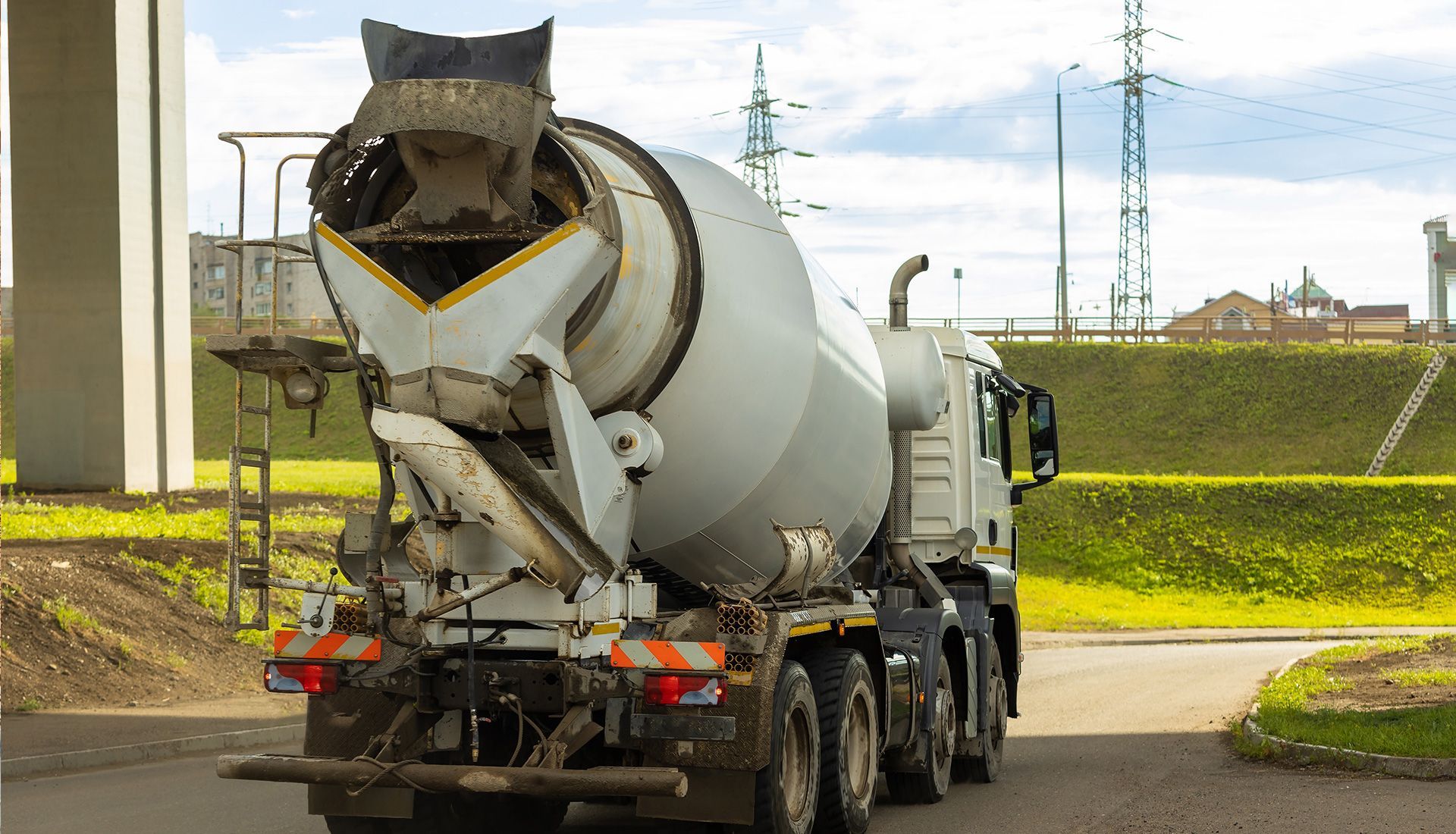 Concrete mixer truck on a road, gray and white with orange safety reflectors, under a bridge near power lines.