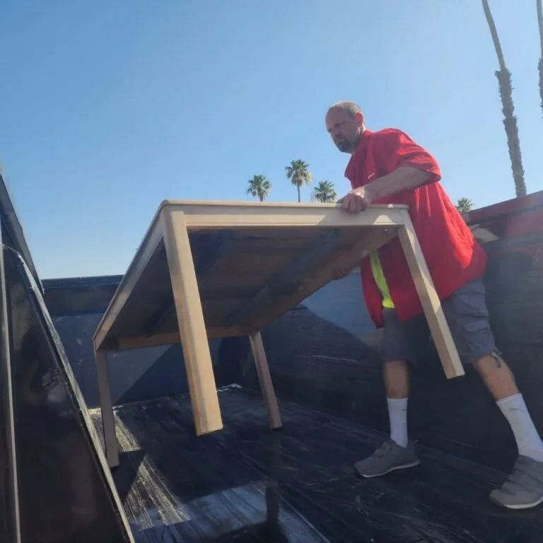 Person in red shirt loading a beige table into a truck bed under a blue sky with palm trees.