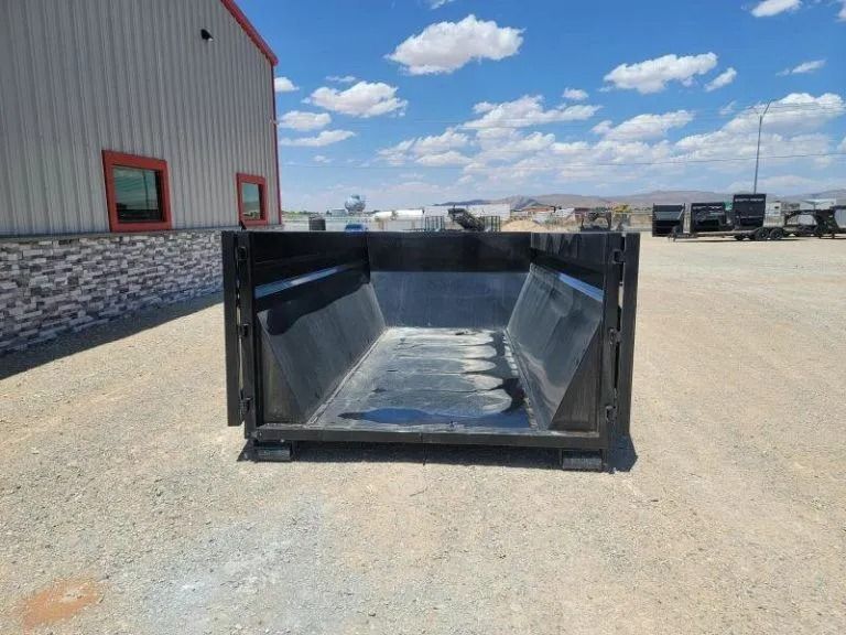 Black dumpster on gravel, in front of a building on a sunny day.