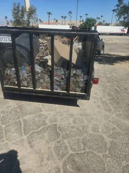 A black trailer filled with trash sits on a cracked pavement parking lot on a sunny day.