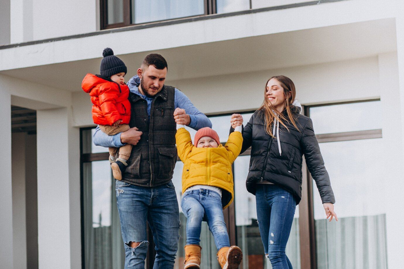Family of four smiling, holding hands, near a modern white house.