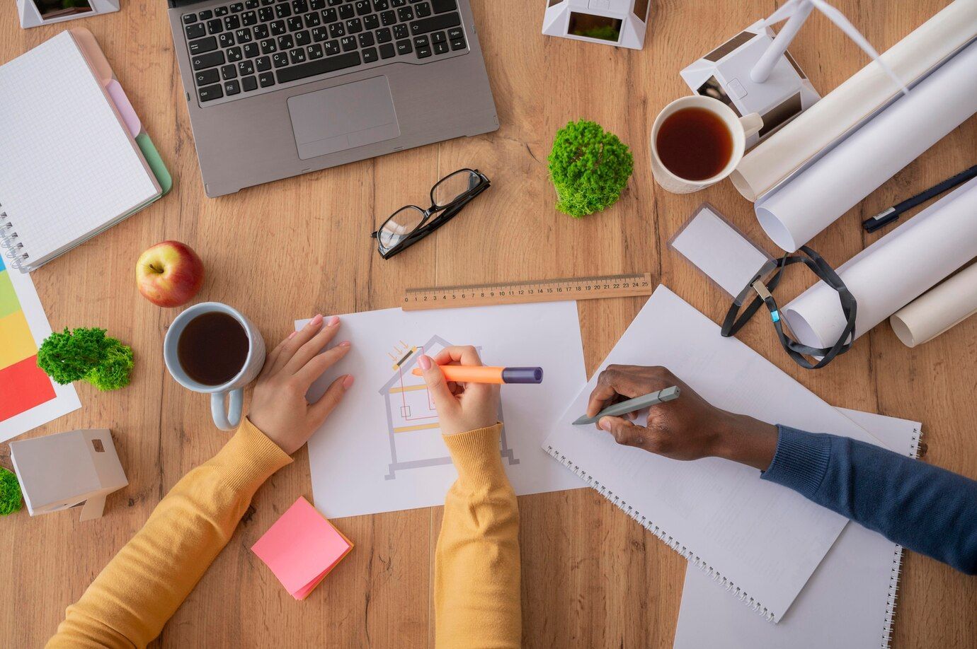 Overhead shot of two people working on architectural drawings at a desk, with laptop, coffee, and supplies.