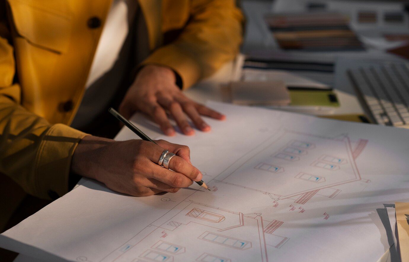 Person in yellow jacket drawing on a blueprint at a desk, illuminated by sunlight.