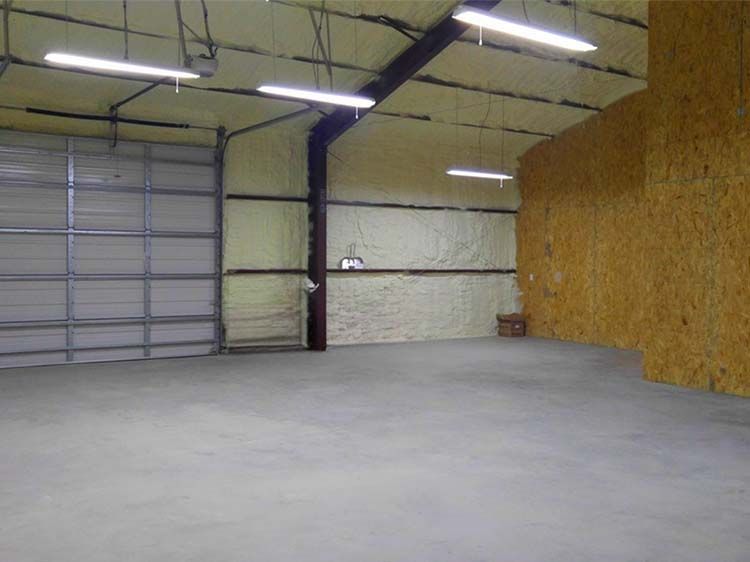 Empty warehouse interior with concrete floor, metal beams, and overhead lights. Garage door on left, plywood wall on right.
