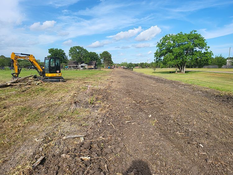 An excavator works on a dirt path in a field under a partly cloudy sky.