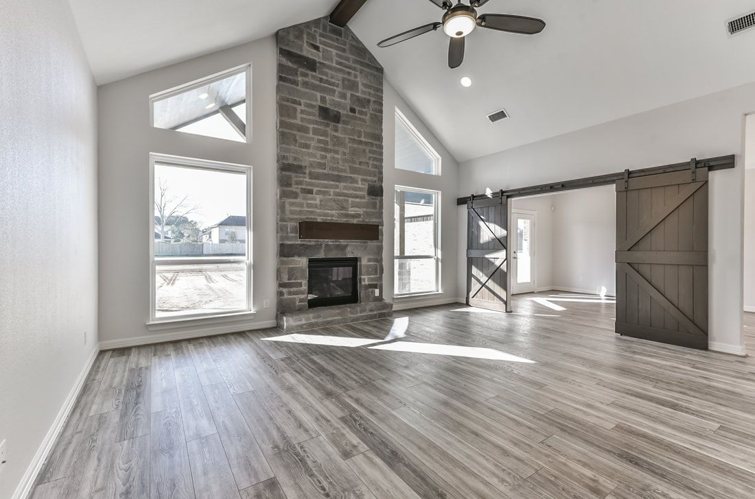 Spacious living room with vaulted ceiling, stone fireplace, large windows, wood floors, and sliding barn door.