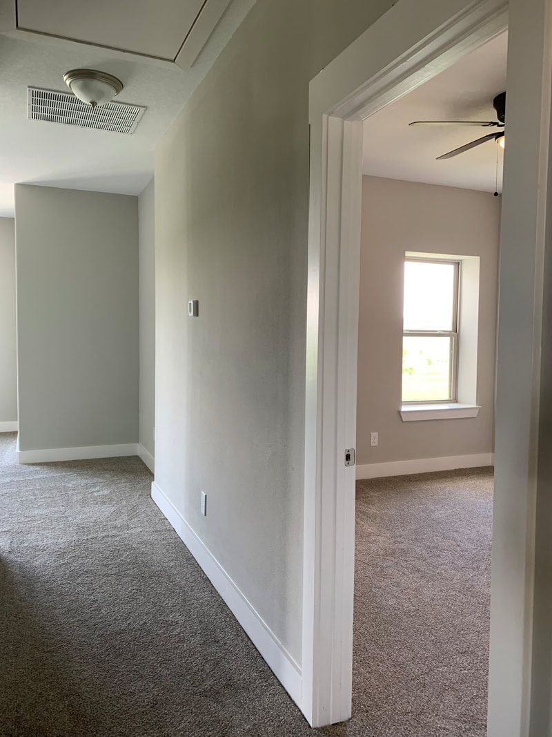 Hallway with gray walls, carpet, and an open doorway to a bedroom with a window.