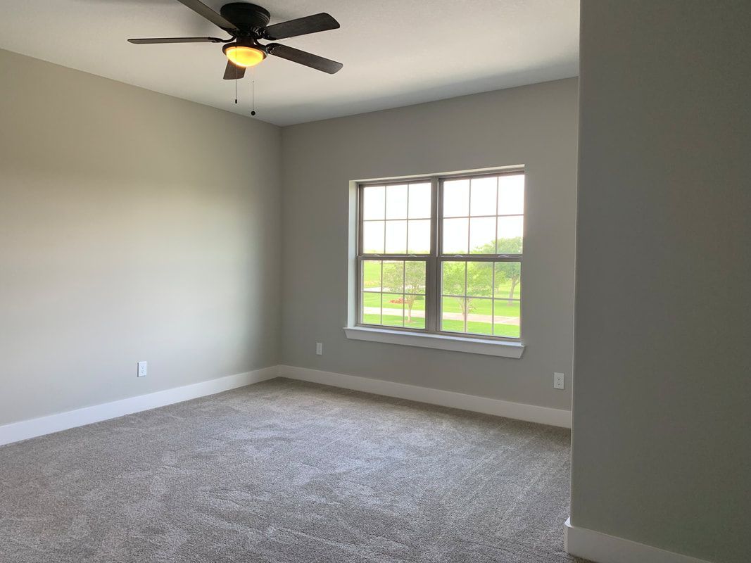 Empty bedroom with light gray walls, carpet, and a window overlooking greenery. Ceiling fan.