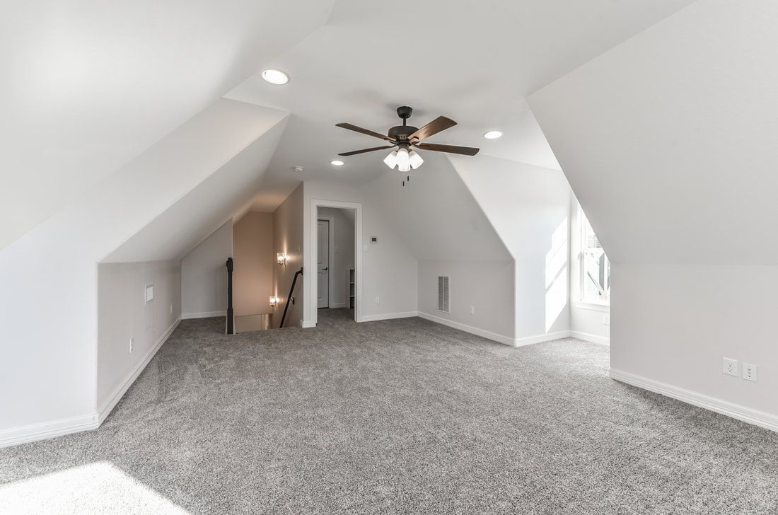 Spacious attic room with gray carpet, angled white walls, ceiling fan, and recessed lighting.