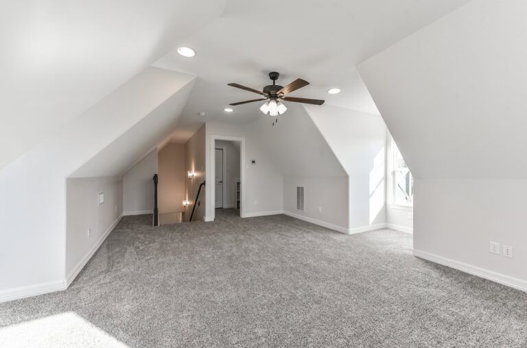 Empty attic room with gray carpet, white walls, angled ceiling, and ceiling fan.