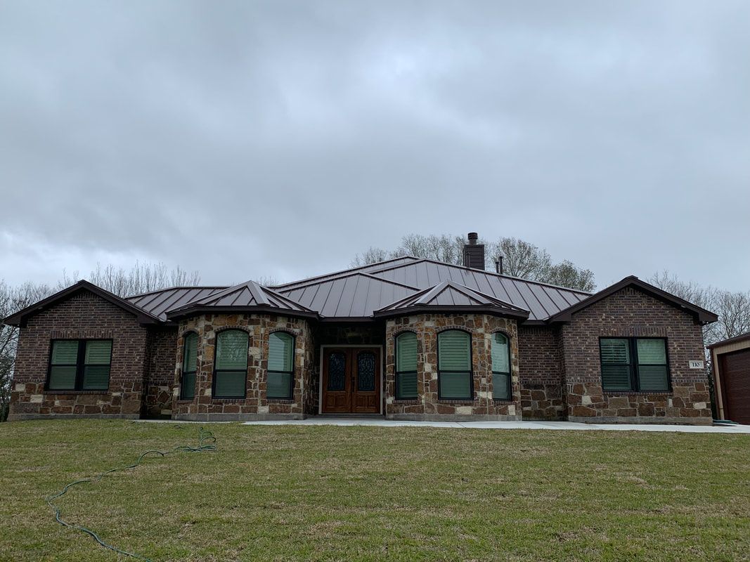 Brown brick and stone house with a dark metal roof under an overcast sky.