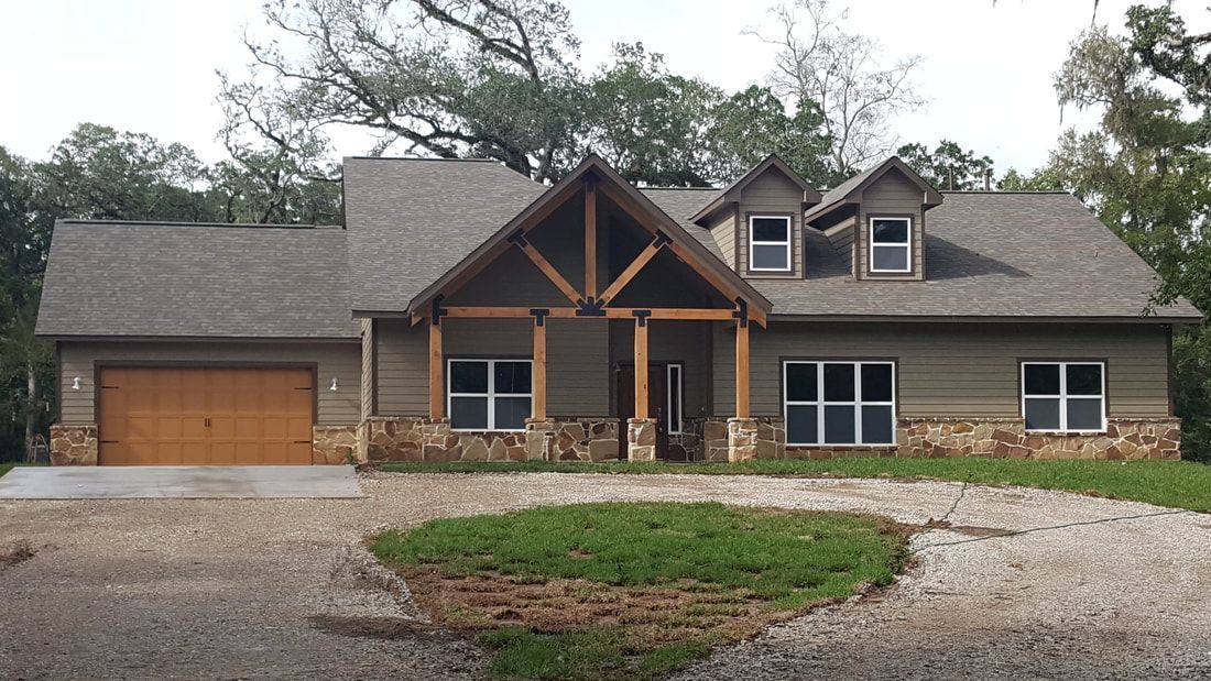 House with brown garage door, stone and wood accents, surrounded by gravel driveway and trees.