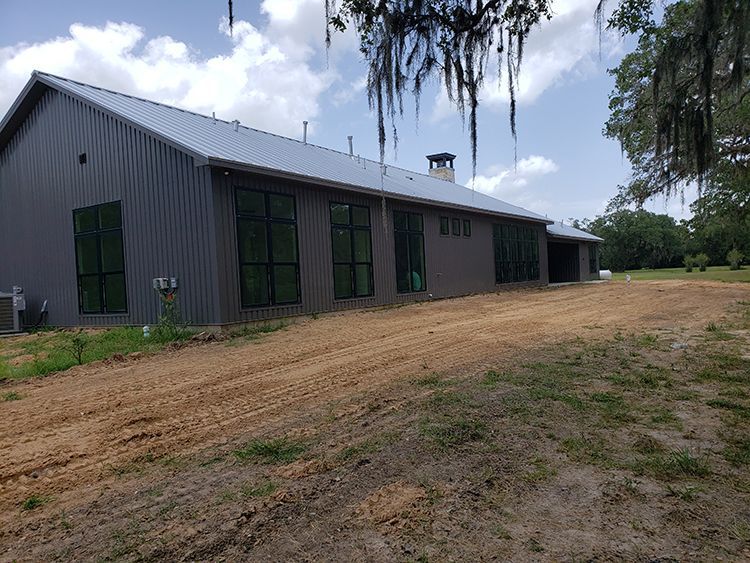 Gray metal building with large windows and a dirt yard on a sunny day.