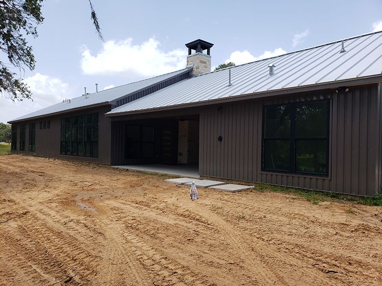 Brown house with large windows, metal roof, and chimney, set on a dirt lot.