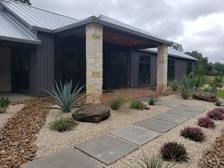 Modern home exterior with stone columns, metal roof, gravel landscaping, and square stepping stones.