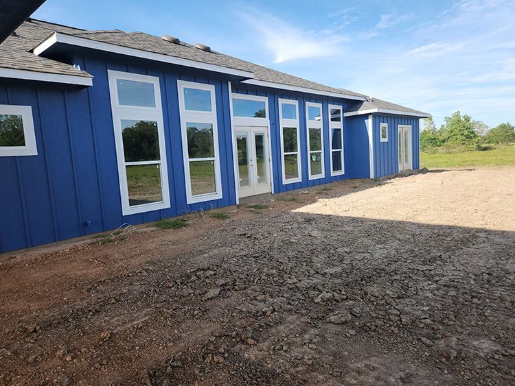 Blue house with white-framed windows, surrounded by dirt and gravel under a blue sky.