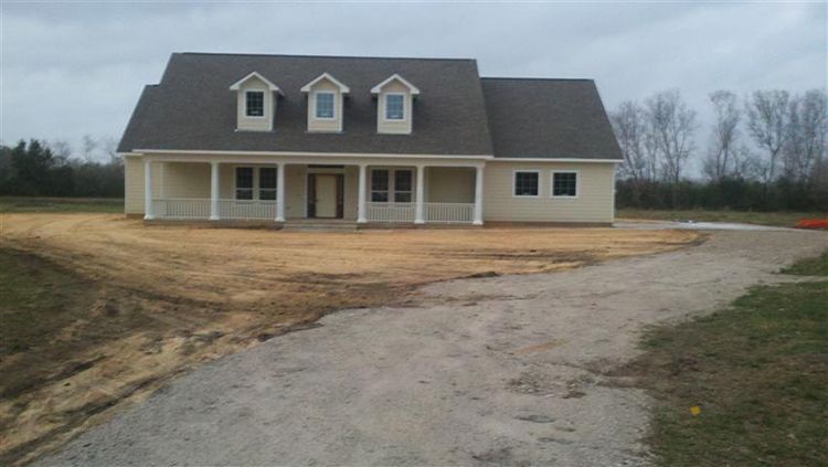 Beige house under construction with porch, dormers, and gravel driveway on a cloudy day.