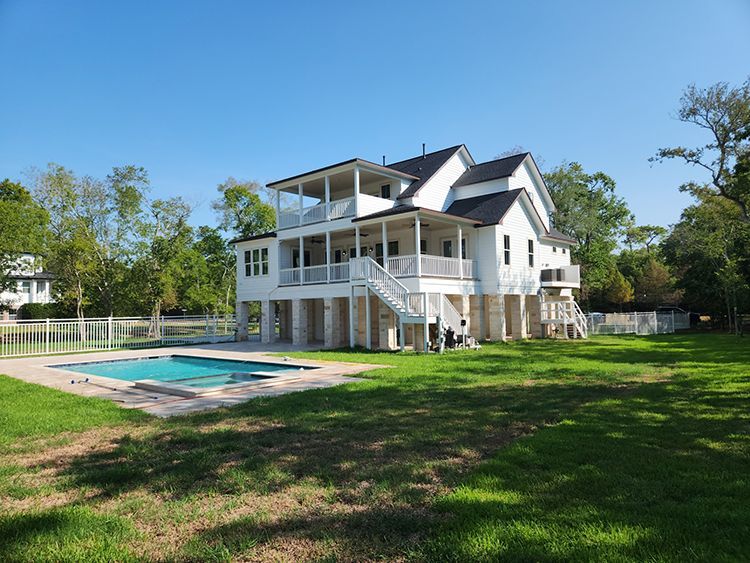 White house with balconies on raised supports, pool in front, green lawn, blue sky.