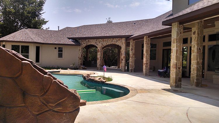 Backyard with a pool, slide, patio, and arched entrance to the house. Brown roof and light-colored stone accents.