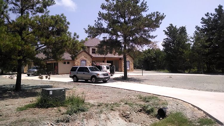 A house with light brown stone and a gray SUV parked in the driveway. Trees and grass surround it.