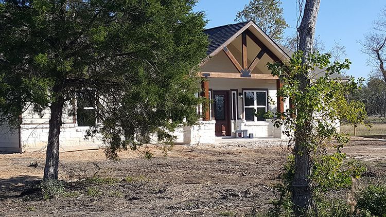 Small, light-colored house with dark roof and brown wood accents. Two windows visible, surrounded by trees.