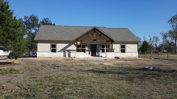 House under construction with light siding, gray roof, and brown porch. Set in a grassy field on a sunny day.