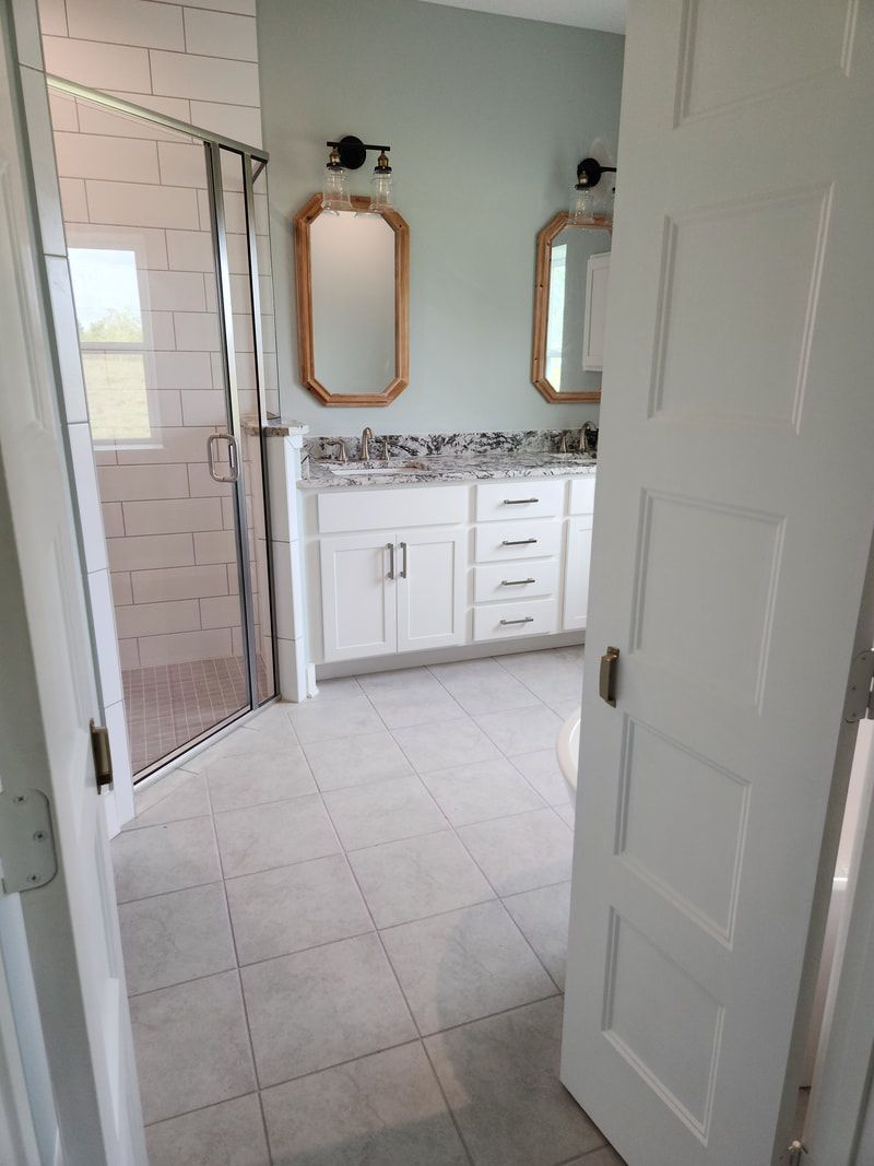 Bathroom with white cabinets, marble countertop, and two framed mirrors. Tile floor and a glass shower.