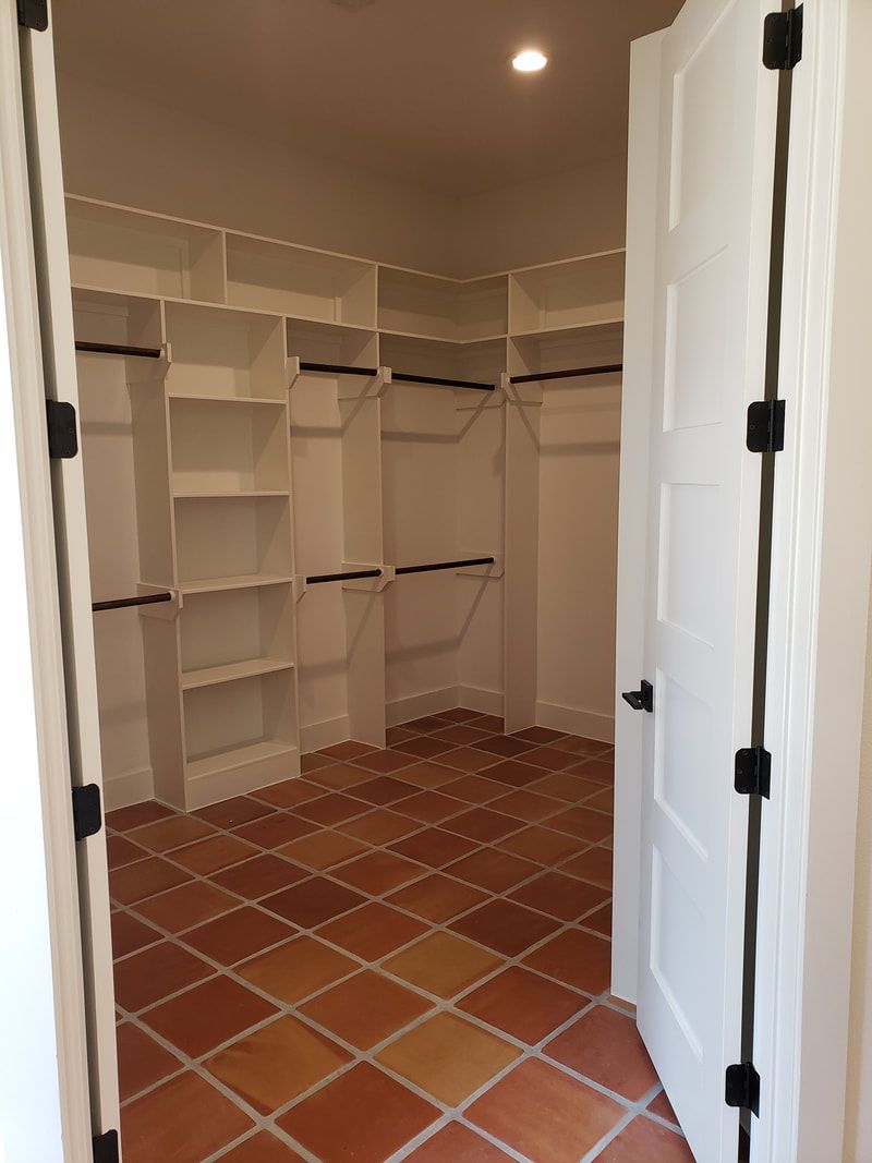 Walk-in closet with white shelving, hanging rods, and terracotta-colored tile floor, viewed through an open white door.