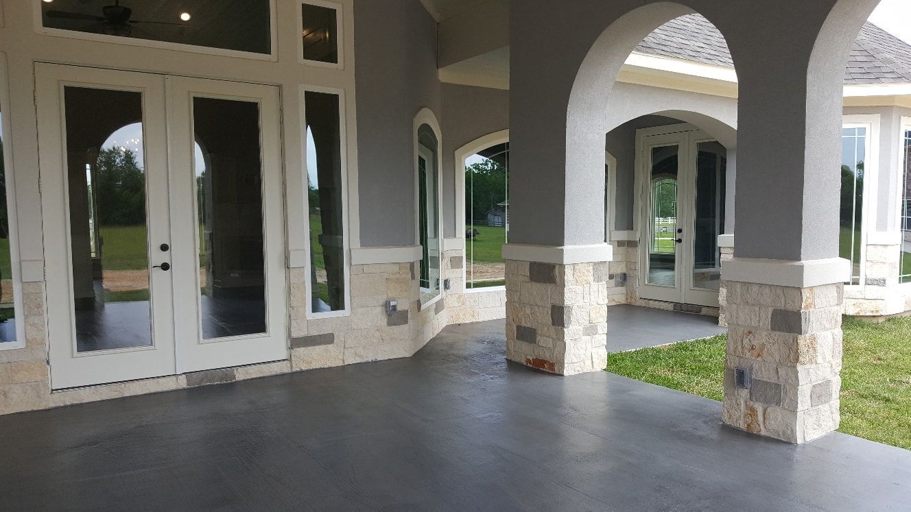 Gray patio with stone columns and arched openings, leading to a house with large windows.