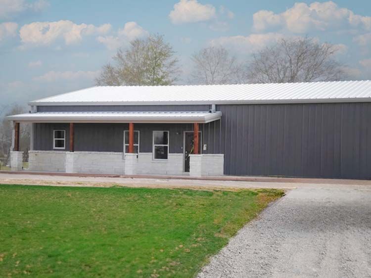 Gray metal building with a white roof and porch, gravel driveway, and green grass.