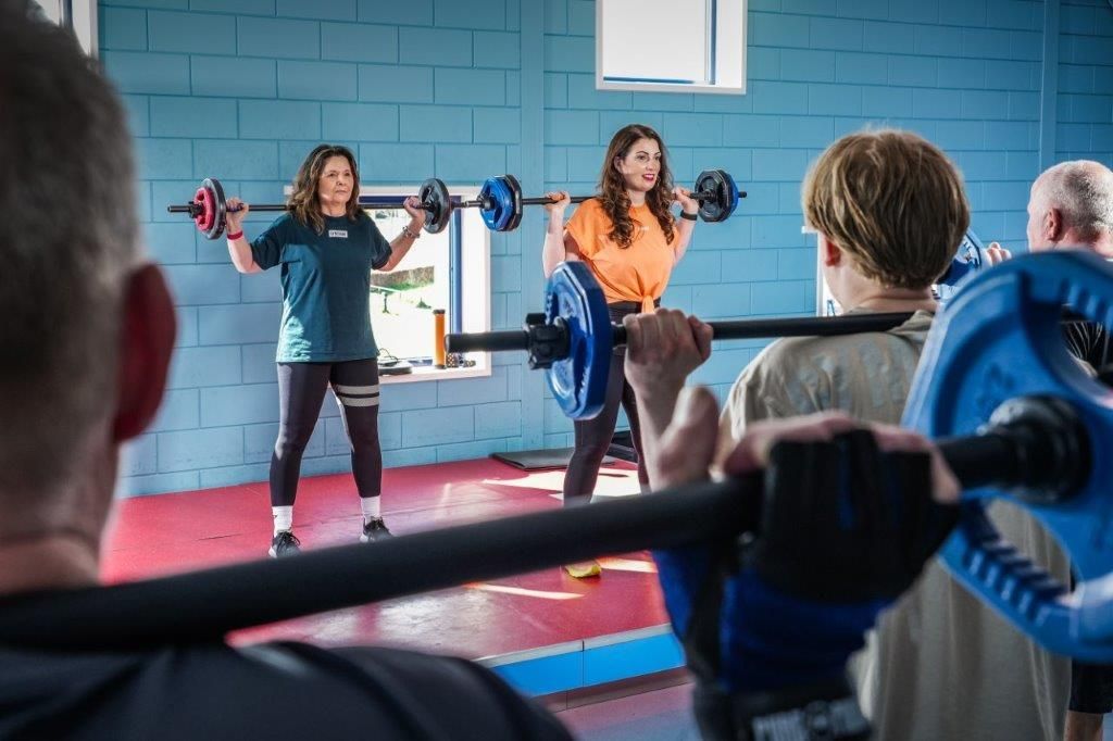 Mensen tillen gewichten in een sportschool. Twee vrouwen op de achtergrond, anderen op de voorgrond.