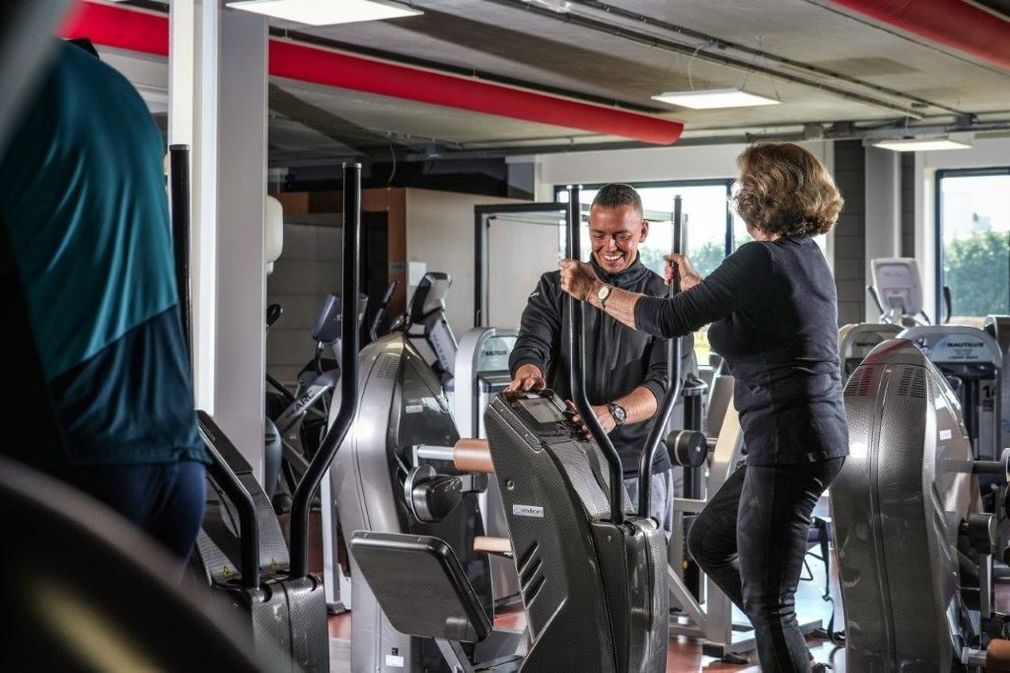 Man en vrouw in de sportschool, op crosstrainers. Man lacht, vrouw fitnesst. De setting is een fitnesscentrum.