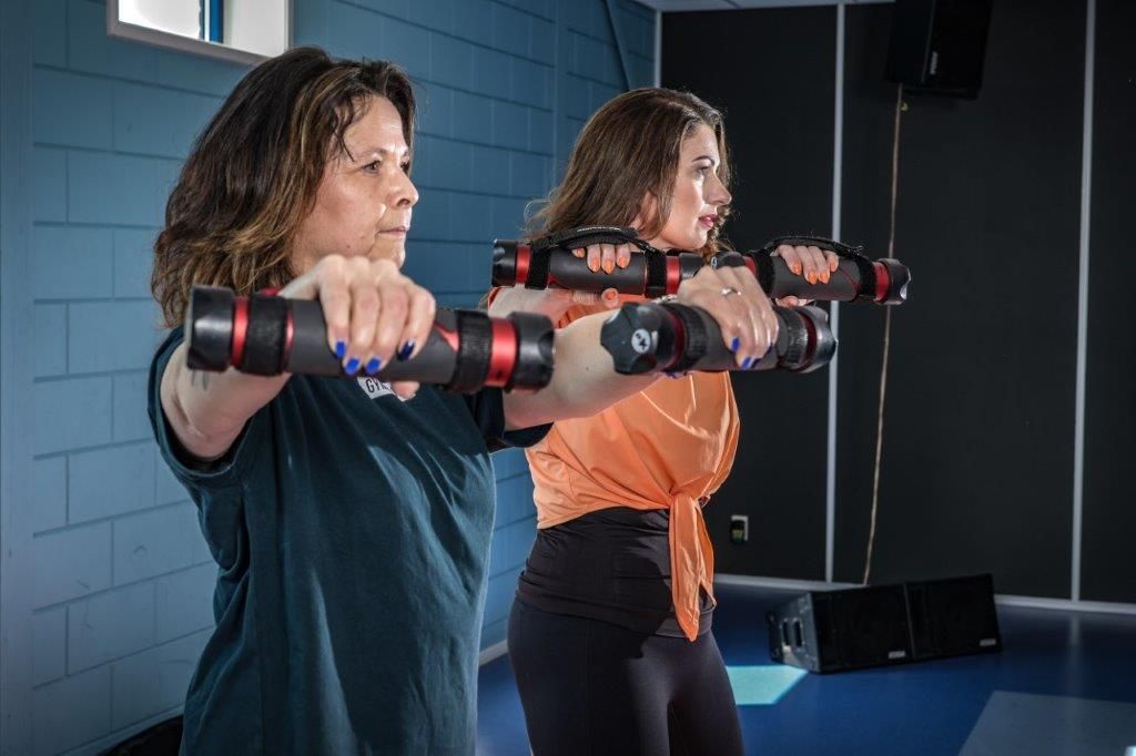 Twee vrouwen oefenen met gewichten in een sportschool. Ze houden de gewichten voor zich omhoog.