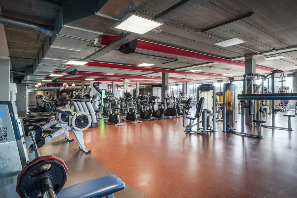 Gym interior with exercise machines on a red and brown floor. Ceiling has red accents and air vents.
