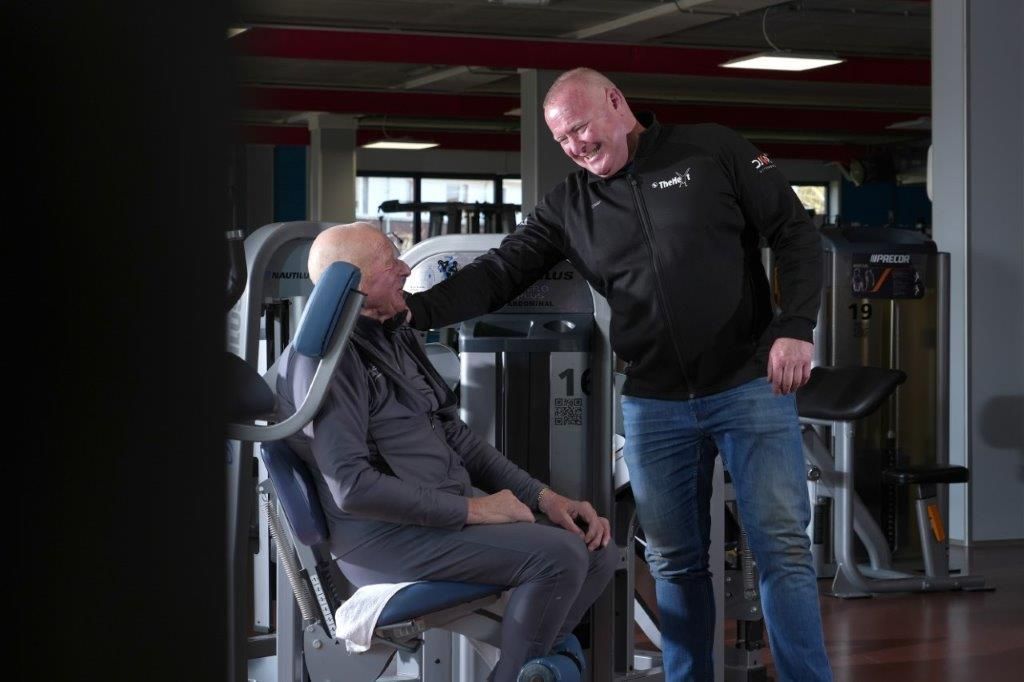 Man in zwart jasje lacht, terwijl hij een andere man helpt met de fitnessapparatuur; interieur van de sportschool.