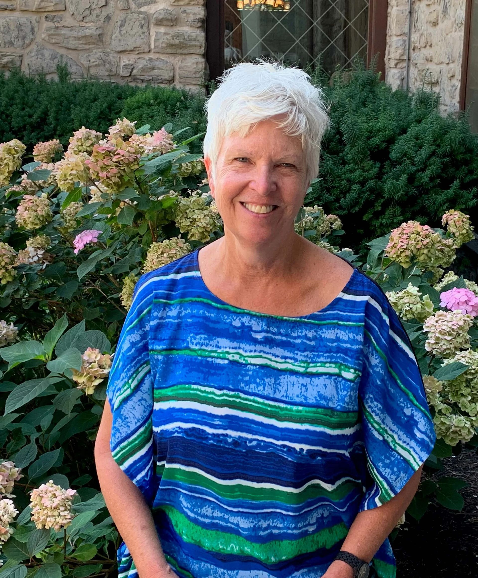 Woman with short white hair smiles, wearing a blue patterned top, in front of flowering bushes.
