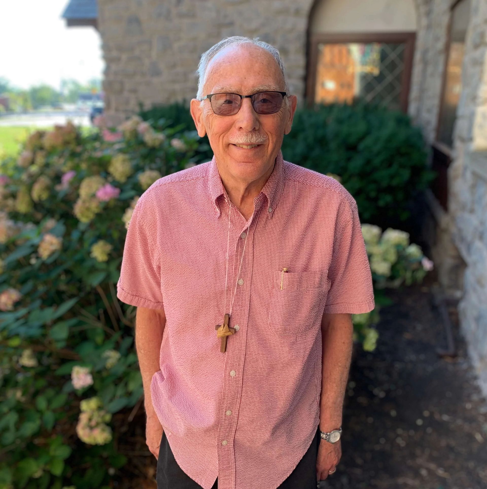 Man wearing glasses and a red patterned shirt stands outside, wearing a cross necklace.
