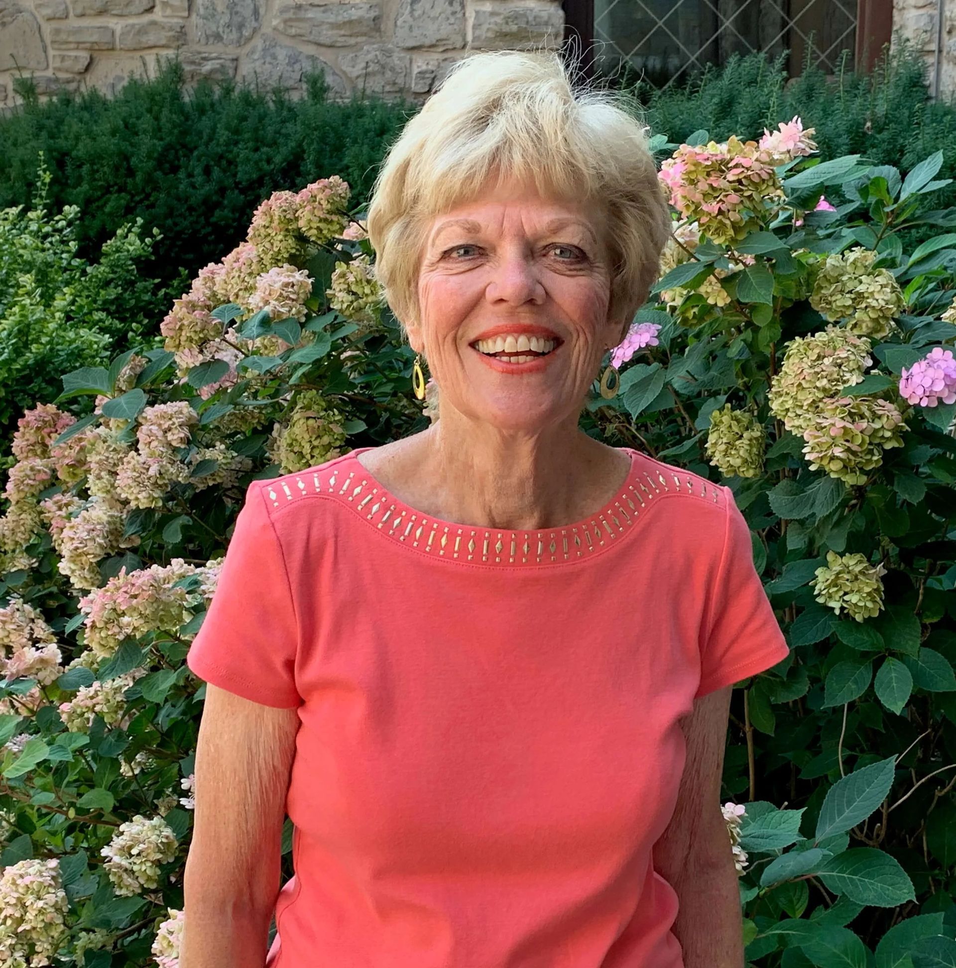 Woman in coral top smiles in front of a blooming hydrangea bush.