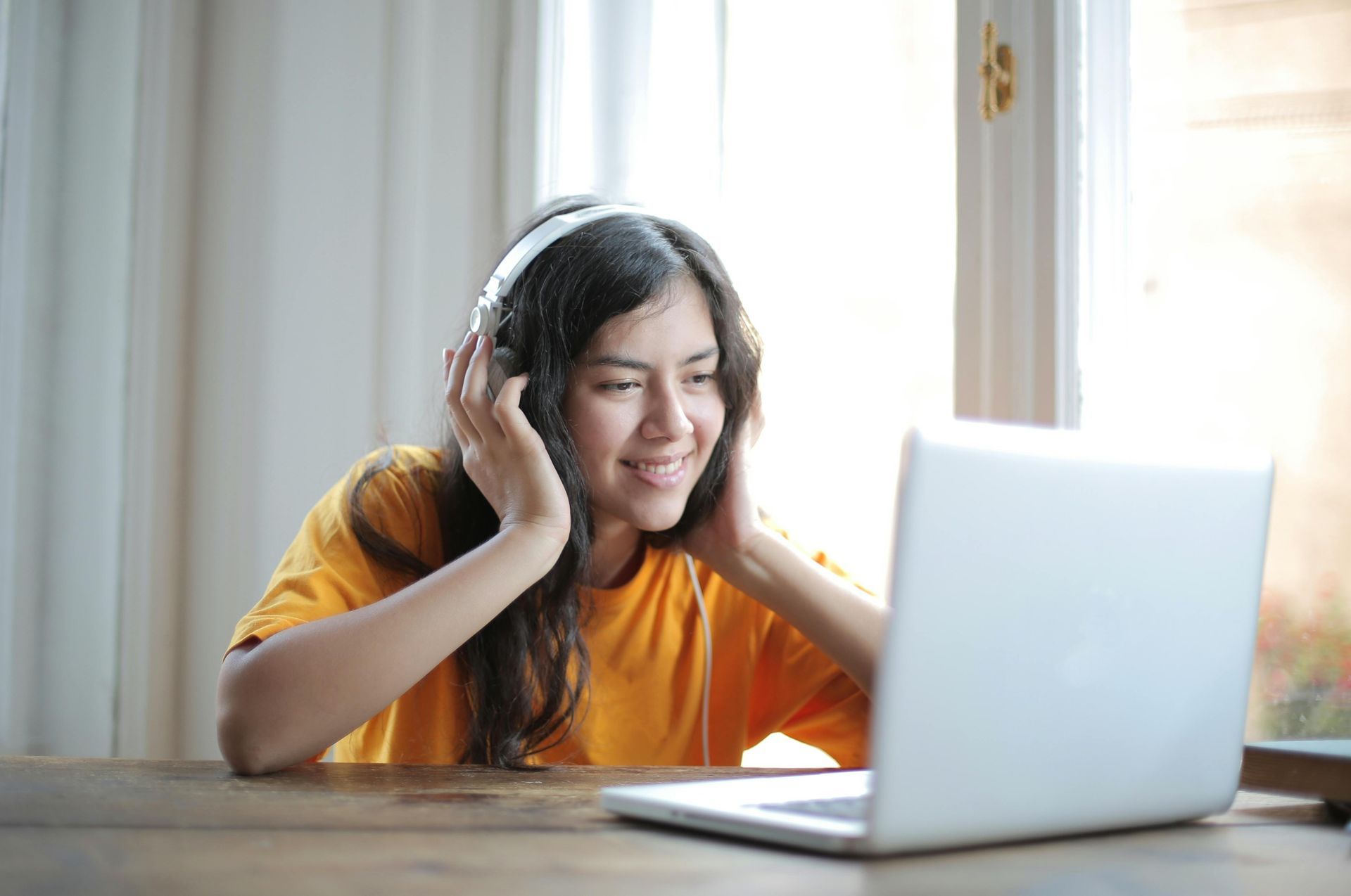Woman in orange shirt wearing headphones smiles at a laptop in front of a window.