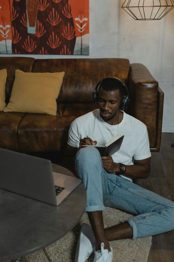 Man wearing headphones writing in notebook, laptop on floor, brown leather couch, abstract art.