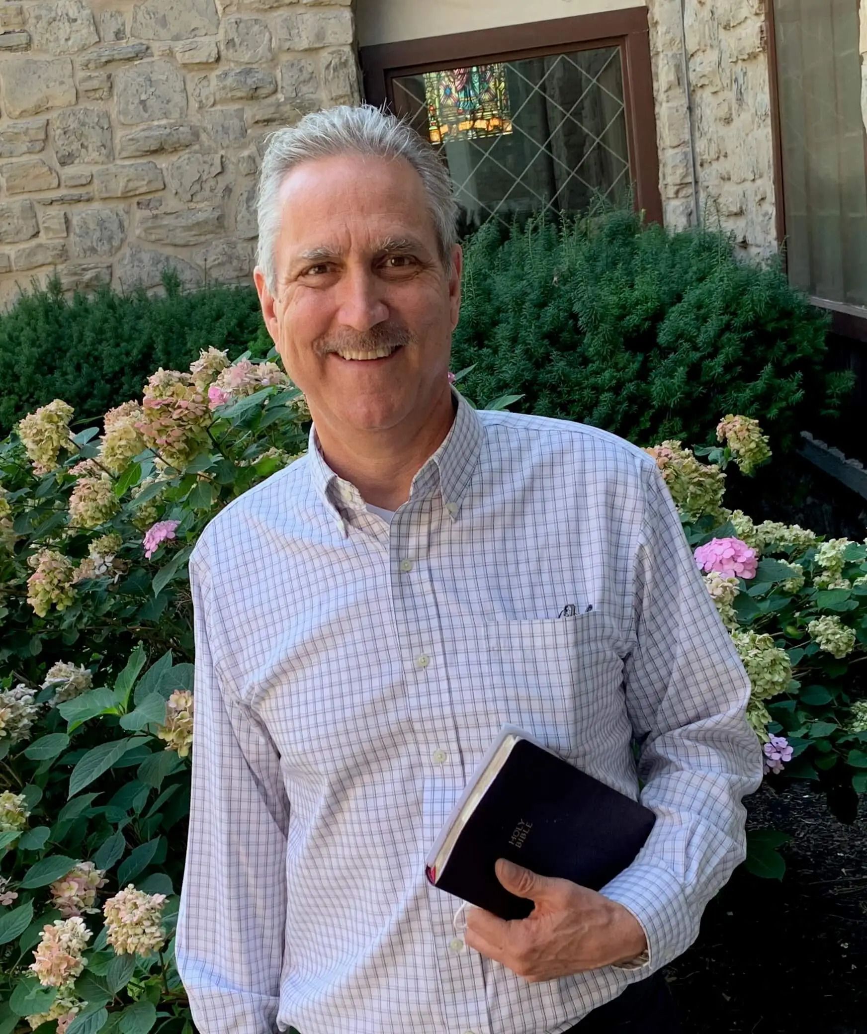 Man in light-colored shirt smiles, holds book, stands in front of stone building and shrubs.