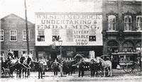 Horse-drawn carriages and people in front of a building with a large sign: 