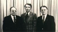 Three men in suits stand in front of a curtain. Center man wears plaid jacket, others dark suits.