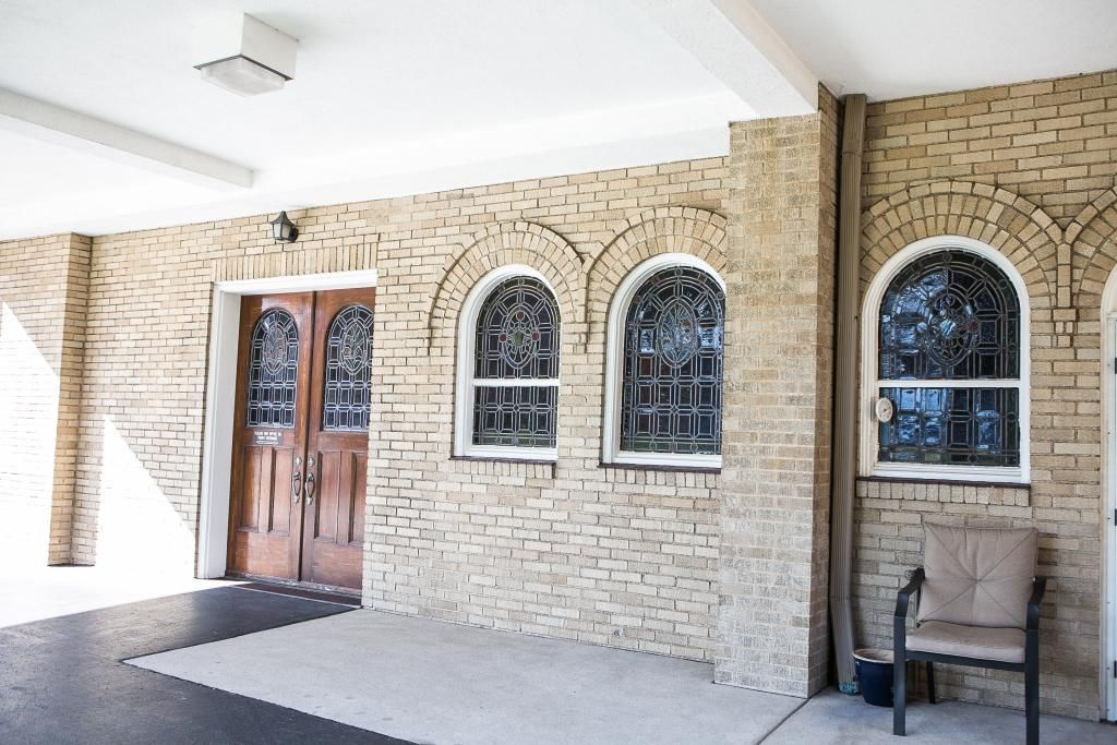 Entrance to a building with arched windows, stained glass, and a wooden door. A chair sits to the side.