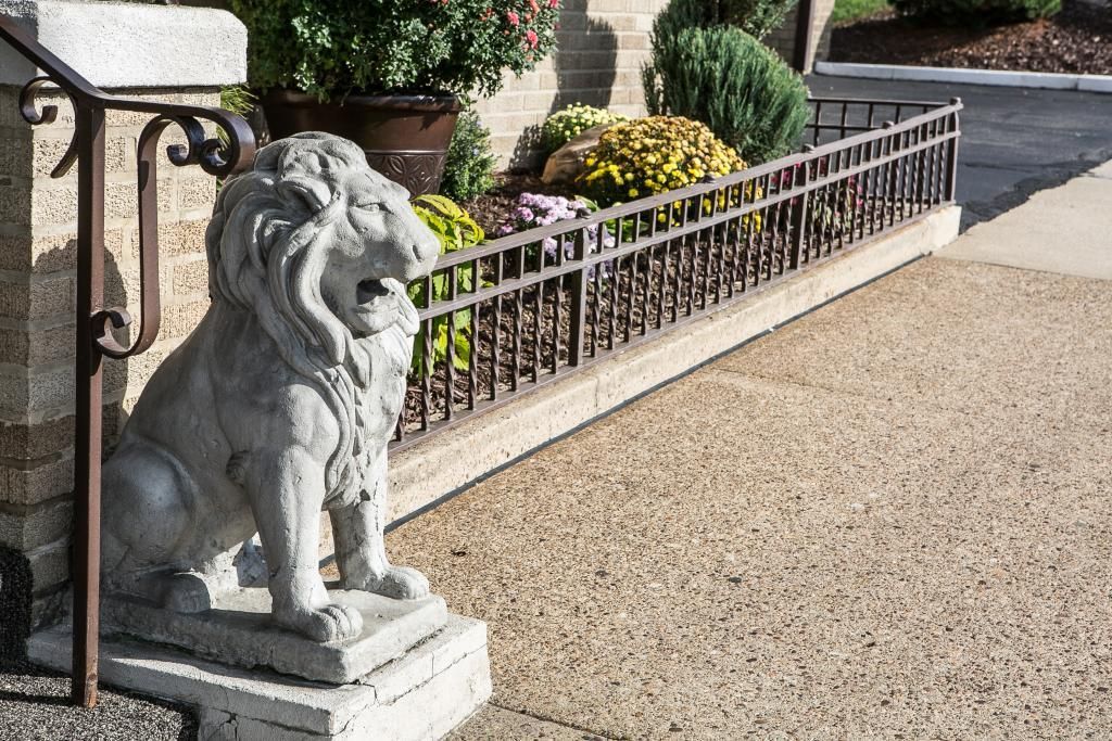 Stone lion statue sits by a flower bed with brown iron railing; sunny outdoor setting.