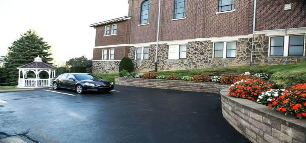 Black car parked near a gazebo and a brick building with a flower bed in front.