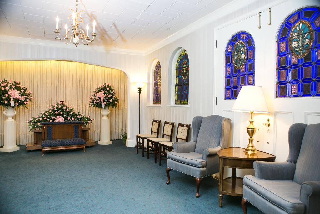 Interior of a funeral home chapel with blue carpet, pews, stained glass windows, and floral arrangements.