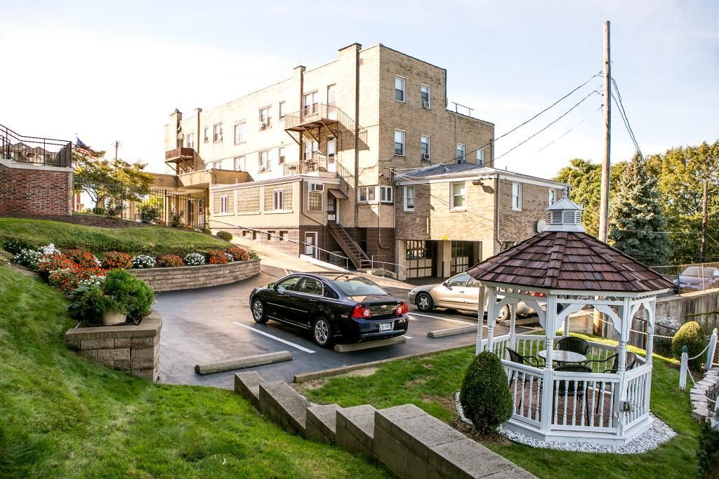 Exterior of a light brown brick building with a gazebo in front. A black car is parked.