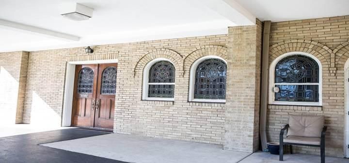 Exterior view of a brick building with arched windows and a wooden door beneath a white overhang. A chair sits to the right.