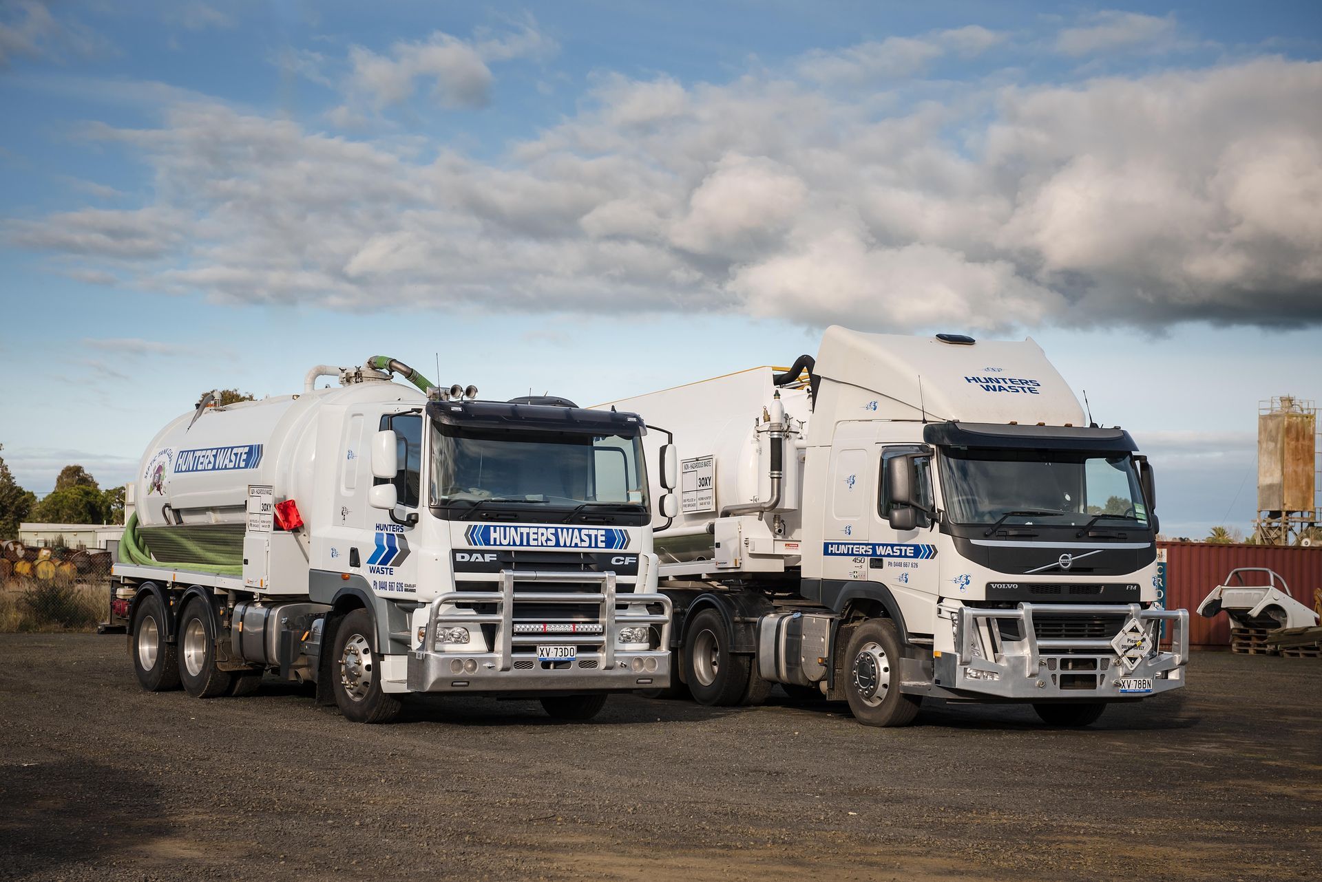 Two white garbage trucks parked side by side on a gravel lot under a cloudy sky Two white garbage trucks parked side by side on a gravel lot under a cloudy sky